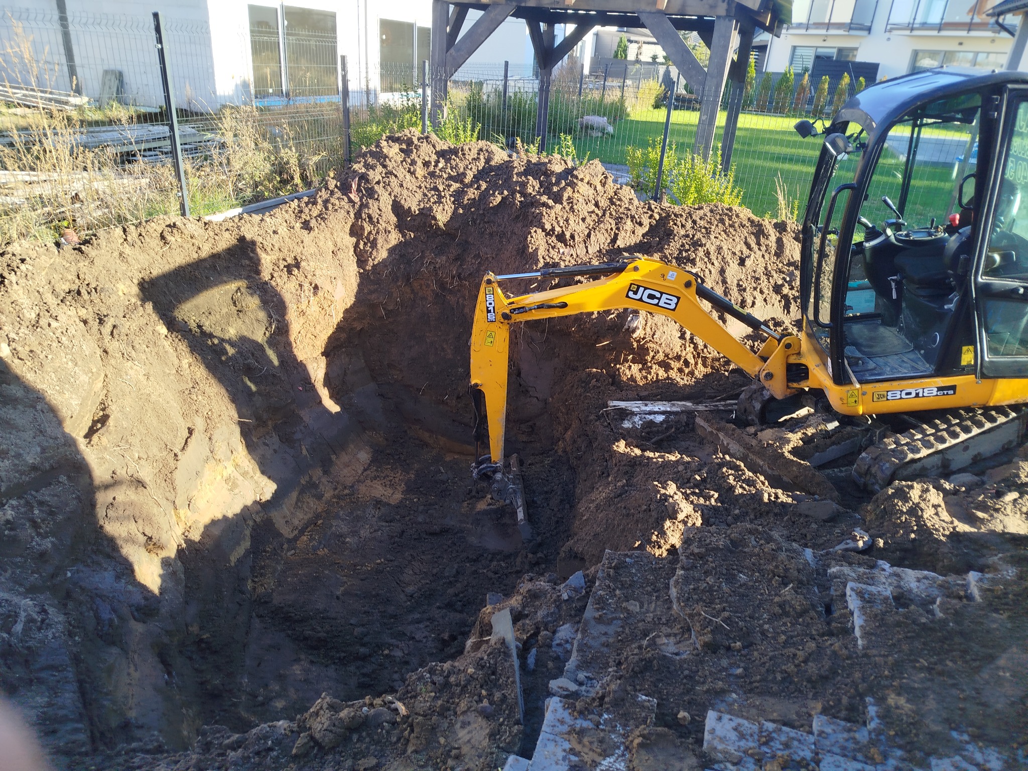 Żółta koparka JCB 3018 CTS w trakcie wykopu ziemi pod fundamenty na działce z widocznym ogrodzeniem i budynkiem w tle, w słoneczny dzień.