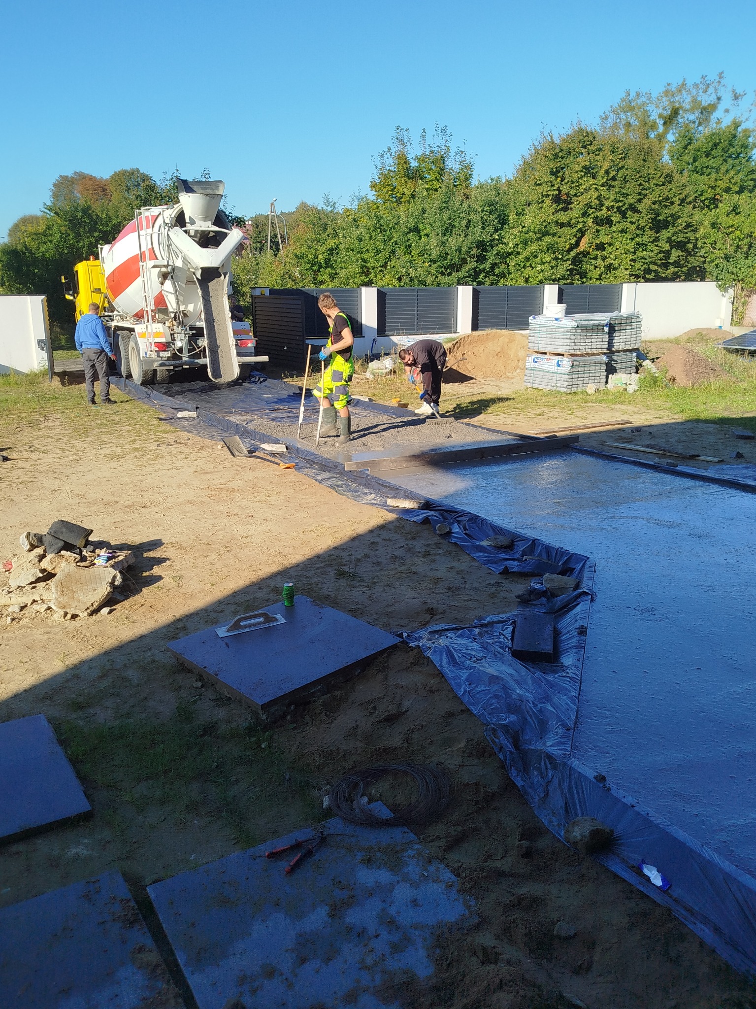 Wylewka betonowa na posesji z użyciem betoniarki, widoczni pracownicy w trakcie rozprowadzania betonu. W tle ogrodzenie panelowe i drzewa.