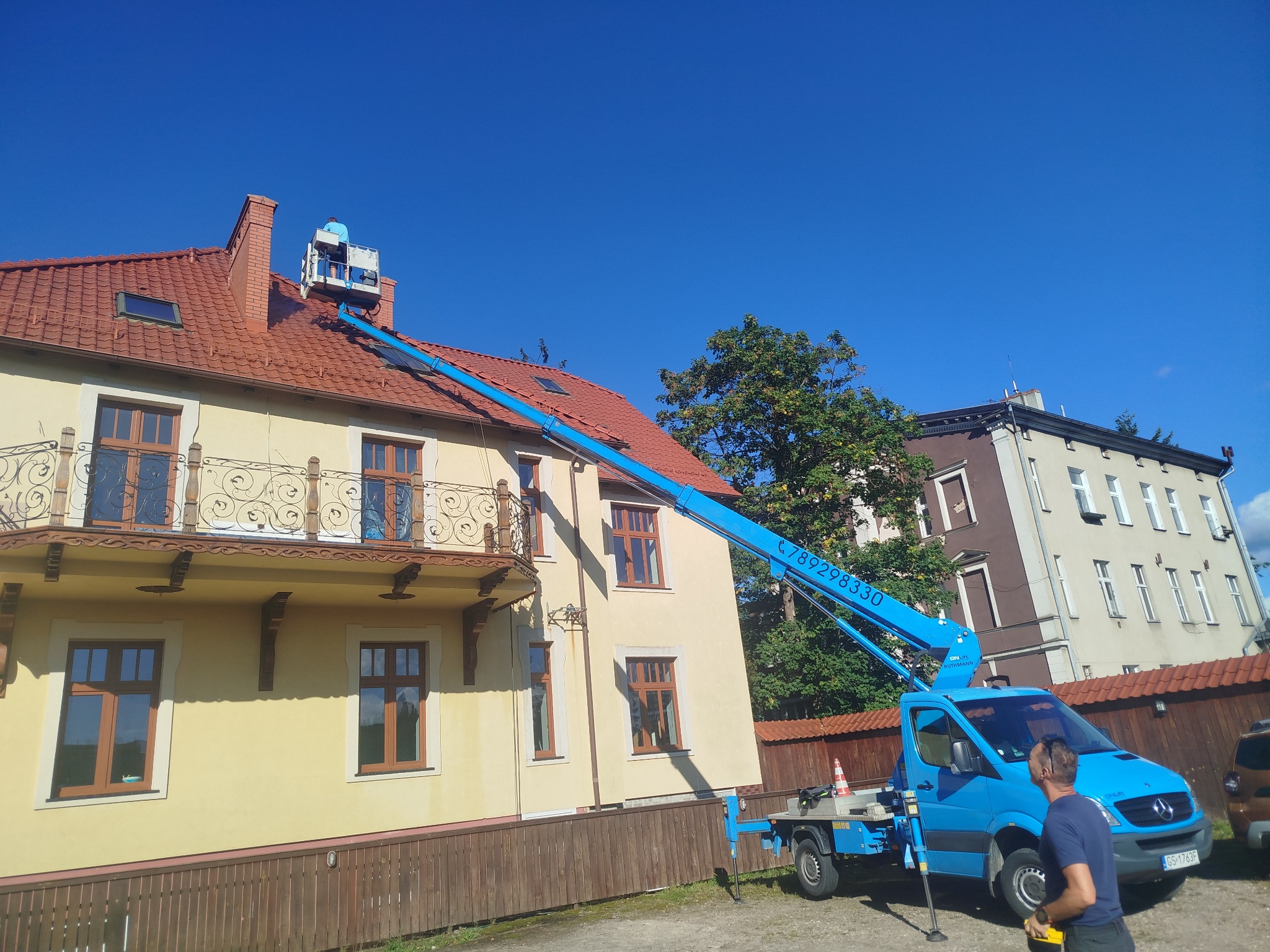 Praca na wysokościach w Gniewino: Błękitny podnośnik koszowy przy budynku z czerwoną dachówką. Osoba w koszu na dachu. Usługi wysokościowe.