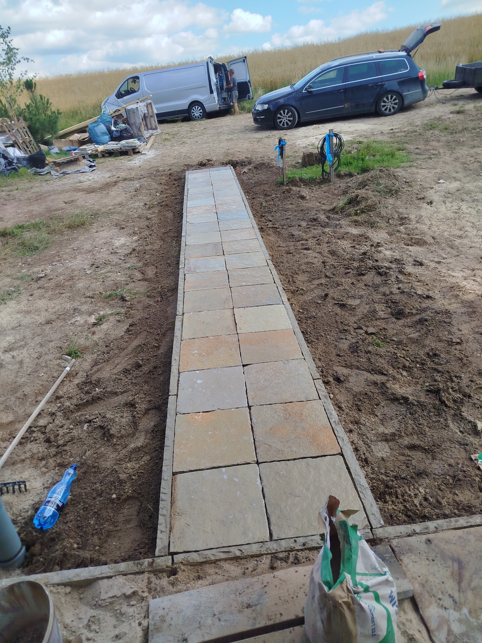 Ścieżka z jasnej kostki brukowej w trakcie układania, widoczne przygotowane podłoże i materiały budowlane, samochody w tle. Ujęcie z bliska, z dołu.