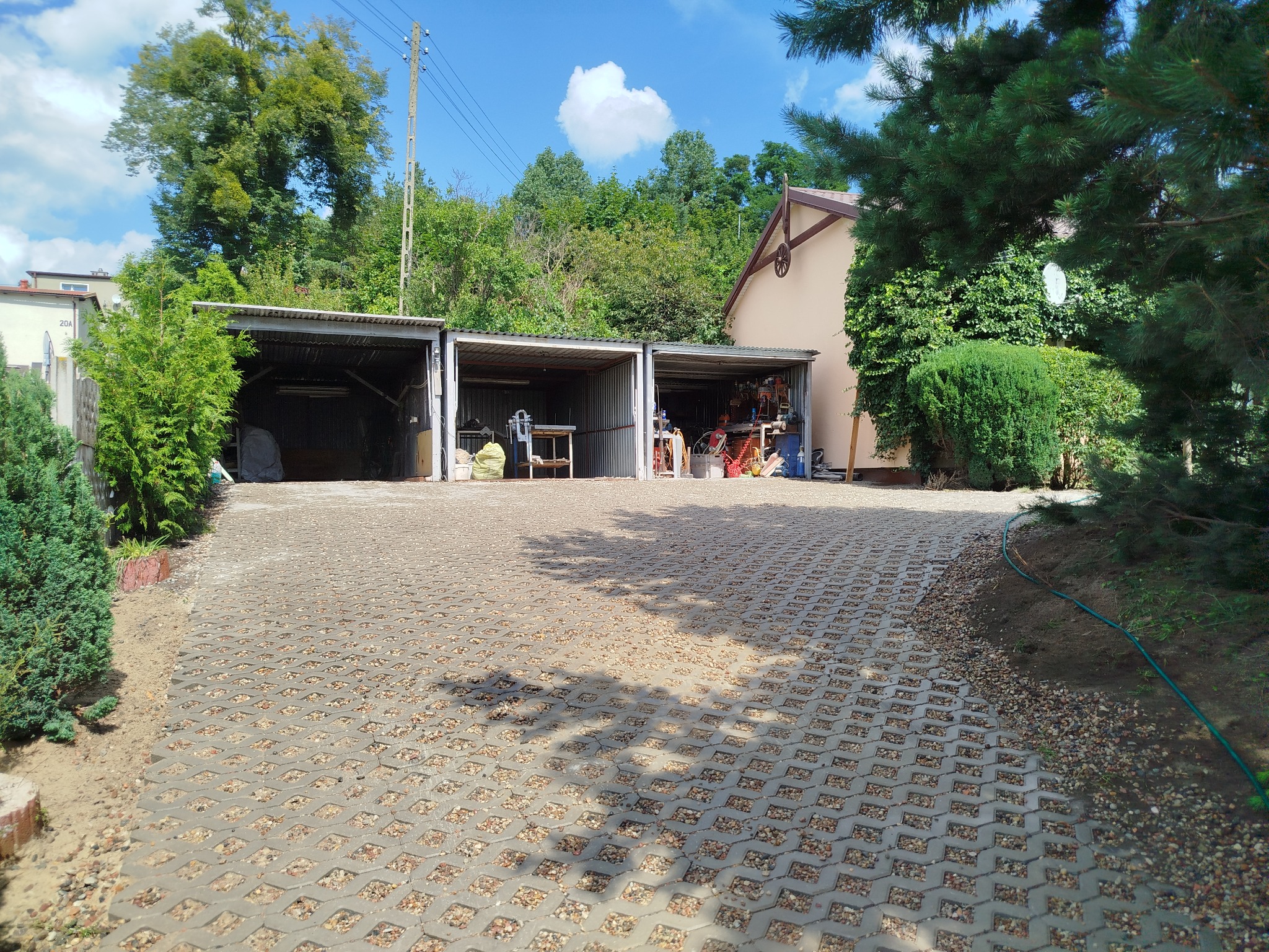 Ułożona szara kostka ażurowa na podjeździe, z widocznymi trzema garażami blaszanymi w tle, otoczona zielenią i drzewami w słoneczny dzień.