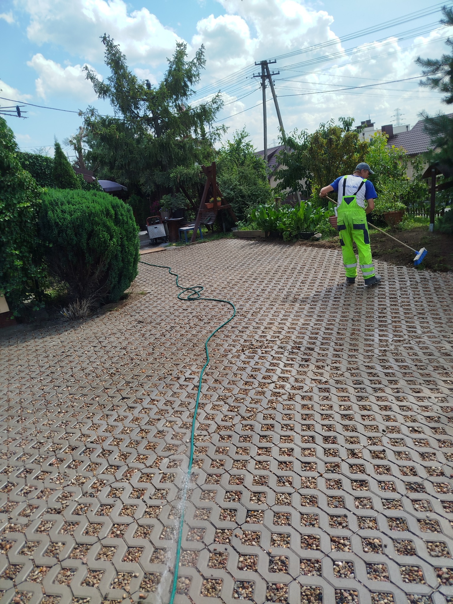 Czyszczenie kostki ażurowej wodą i szczotką przez pracownika w jaskrawozielonym kombinezonie. Wąż ogrodowy na pierwszym planie. Tło: zieleń ogrodu.