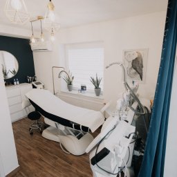 Gabinety Medikos - Jasne wnętrze gabinetu z łóżkiem zabiegowym, nowoczesnym sprzętem i dekoracyjnymi lampami. Minimalistyczny design z akcentami złota i granatu. Widok z perspektywy klienta.