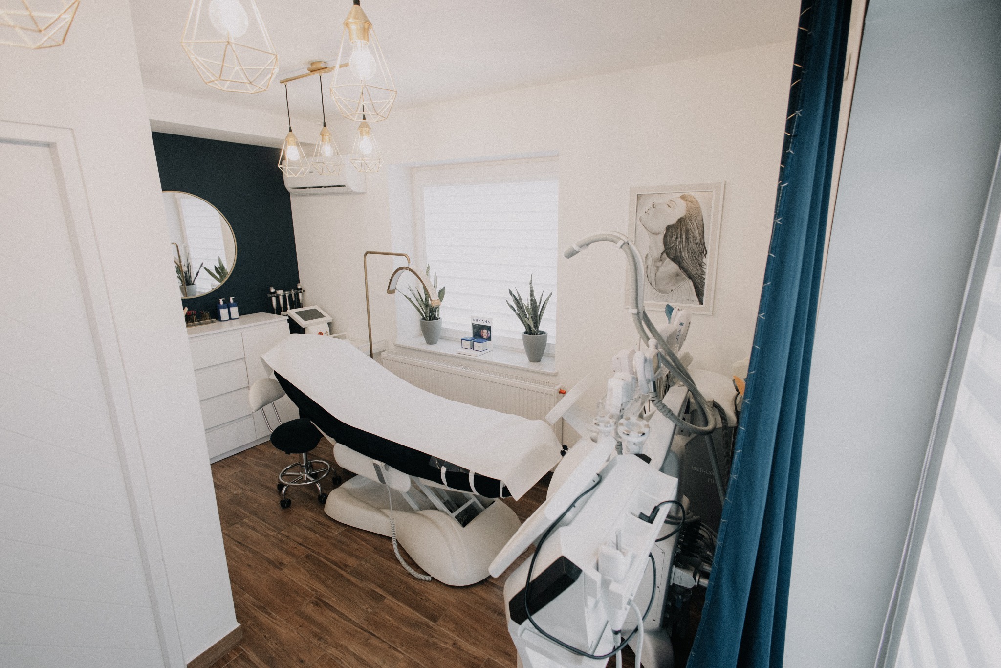 Jasne wnętrze gabinetu z łóżkiem zabiegowym, nowoczesnym sprzętem i dekoracyjnymi lampami. Minimalistyczny design z akcentami złota i granatu. Widok z perspektywy klienta.