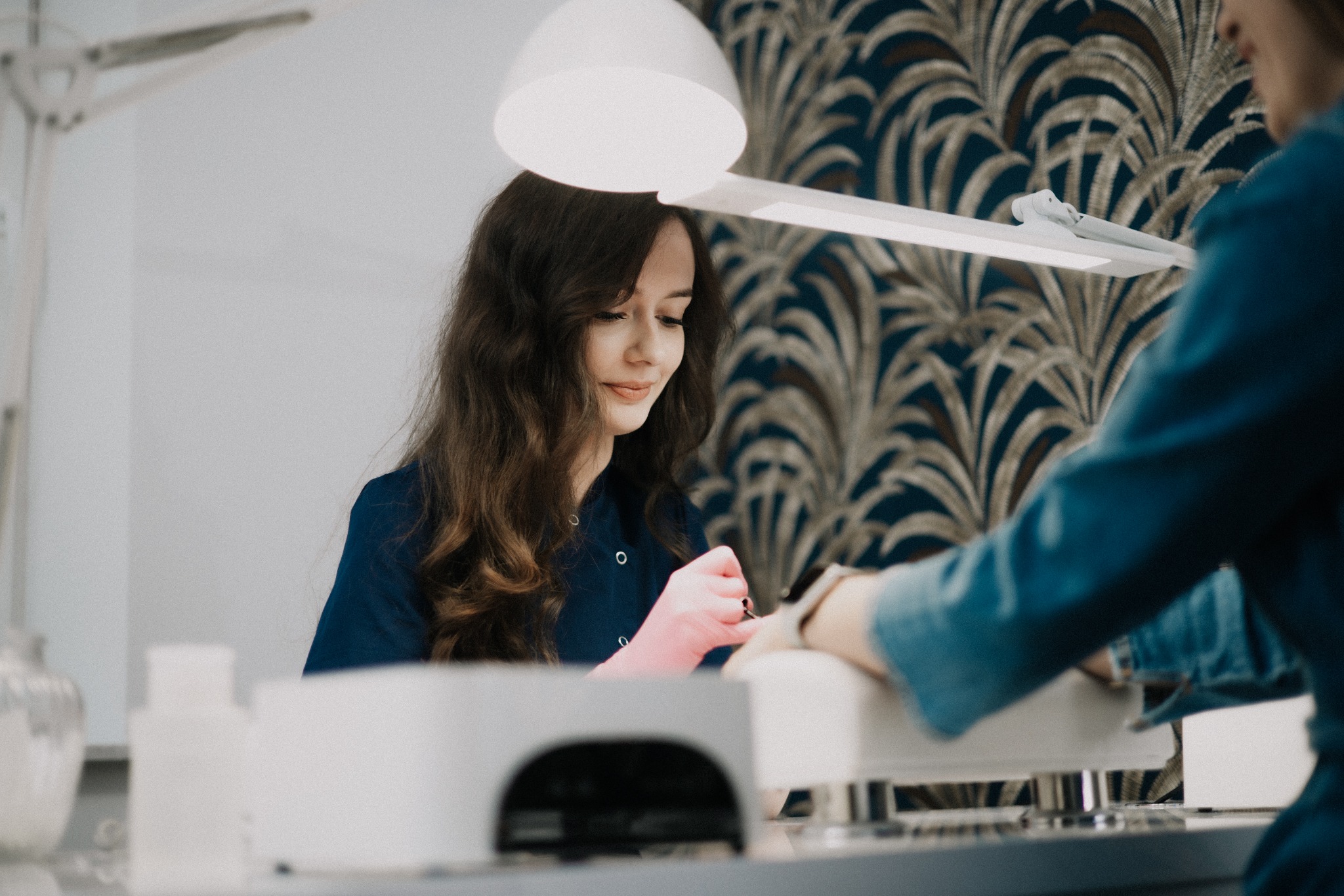 Stylistka paznokci w trakcie pracy nad manicure, używa lampy UV. W tle wzorzysta tapeta i butelki z preparatami. Ujęcie z bliska, ciepłe oświetlenie.