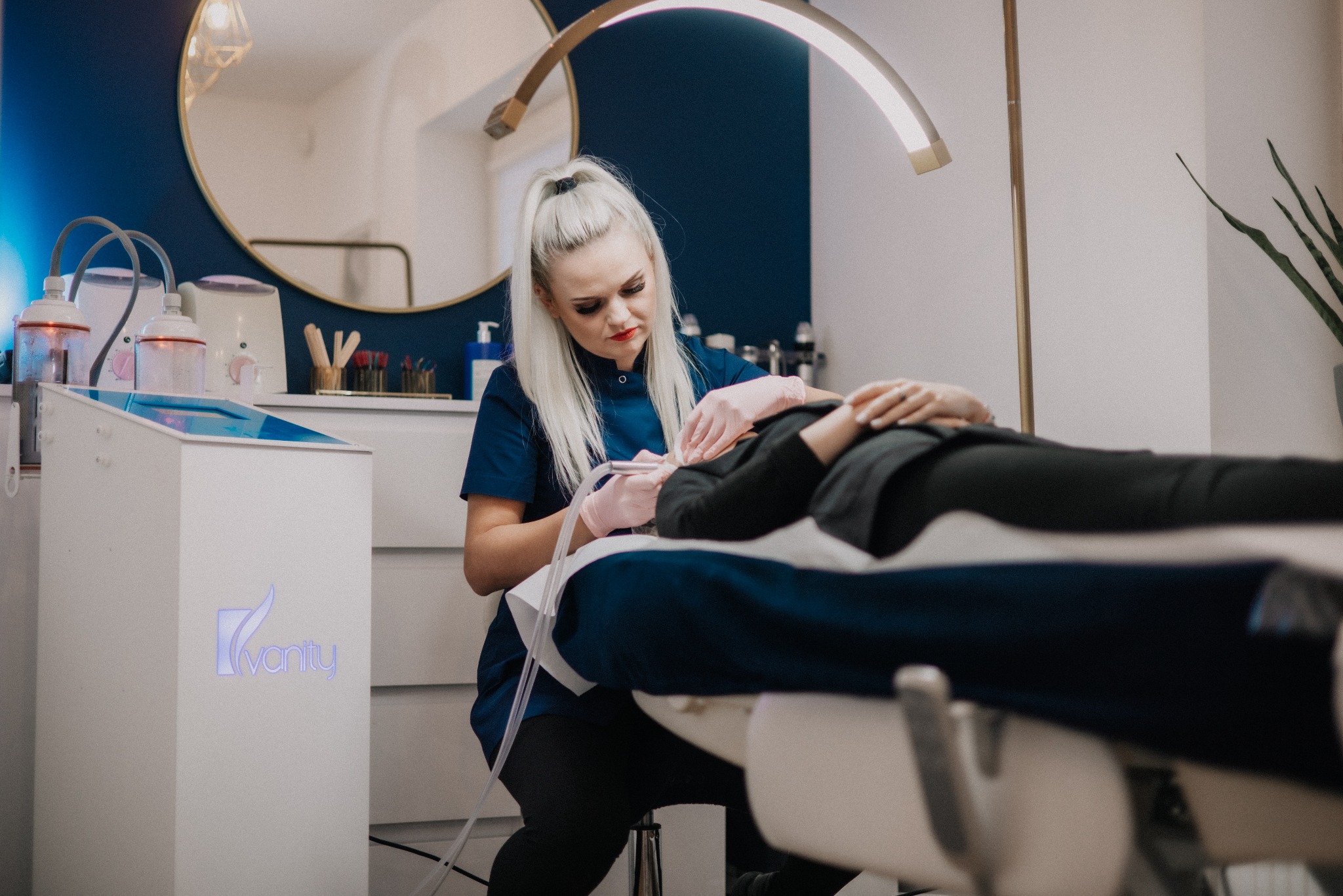 Kosmetolog w niebieskim uniformie wykonuje zabieg na twarzy klientki leżącej na białym fotelu w jasnym gabinecie. Widoczna lampa, lustro i urządzenie kosmetyczne.
