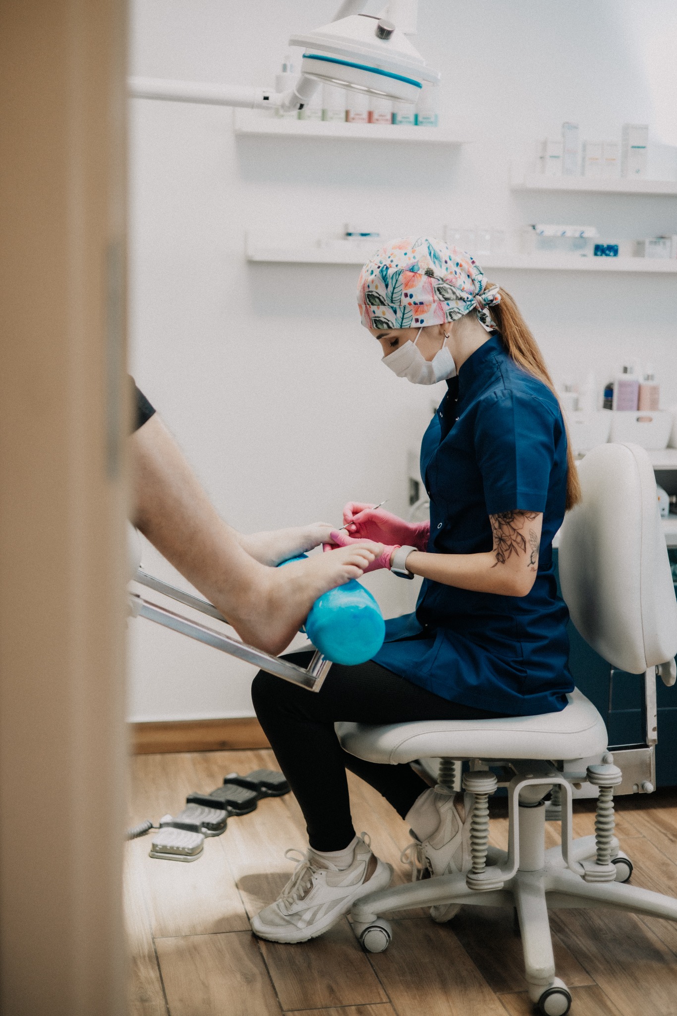 Pedicure w gabinecie: Klient siedzi na fotelu, a pedikiurzystka w masce i rękawiczkach wykonuje zabieg na stopach. Widoczne narzędzia, lampa i półki z kosmetykami.