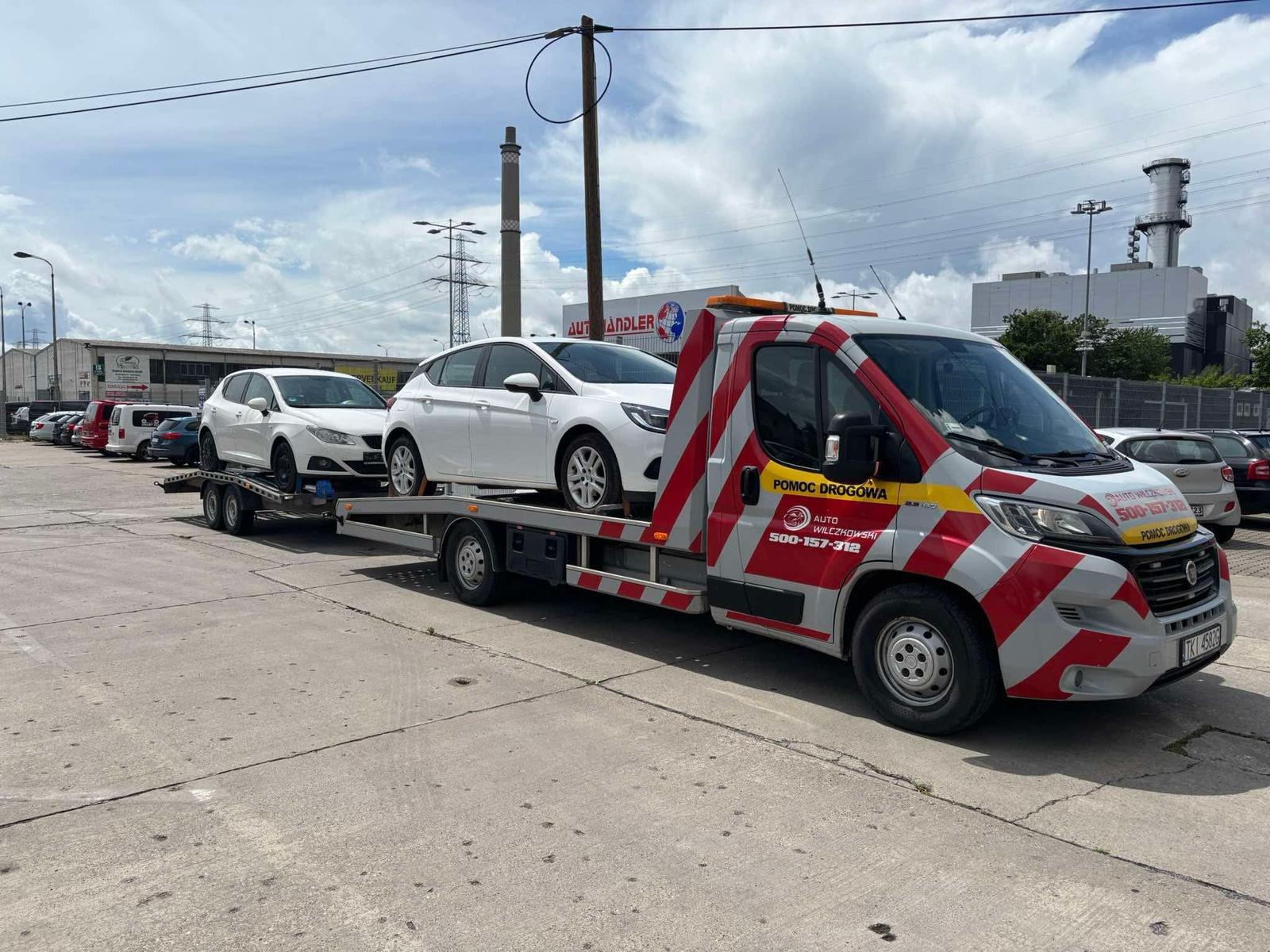 Laweta z dwoma białymi samochodami na platformie, zaparkowana na betonowym placu. W tle budynki i słupy energetyczne. Na lawecie widoczne logo firmy 'Auto Wilczkowski'.