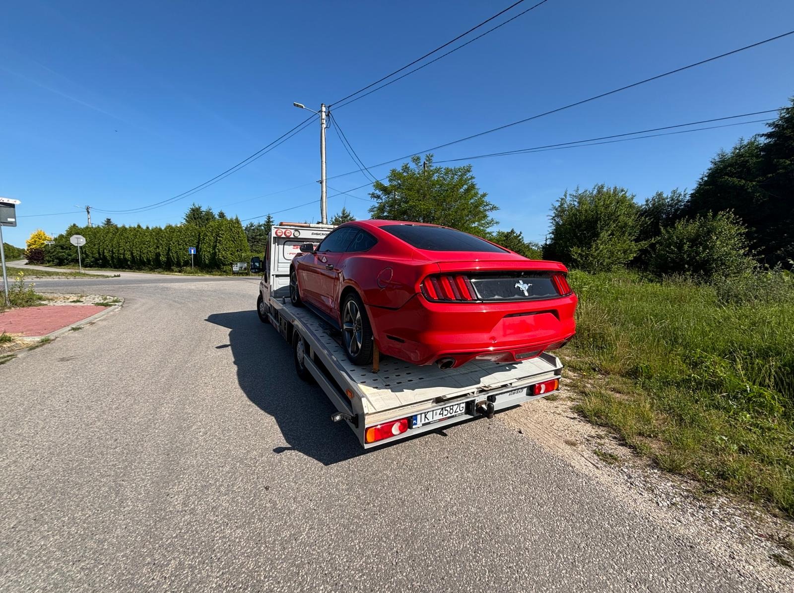 Czerwony Ford Mustang na lawecie, widok z tyłu, tablica rejestracyjna widoczna, słoneczny dzień, asfaltowa droga, zieleń w tle.