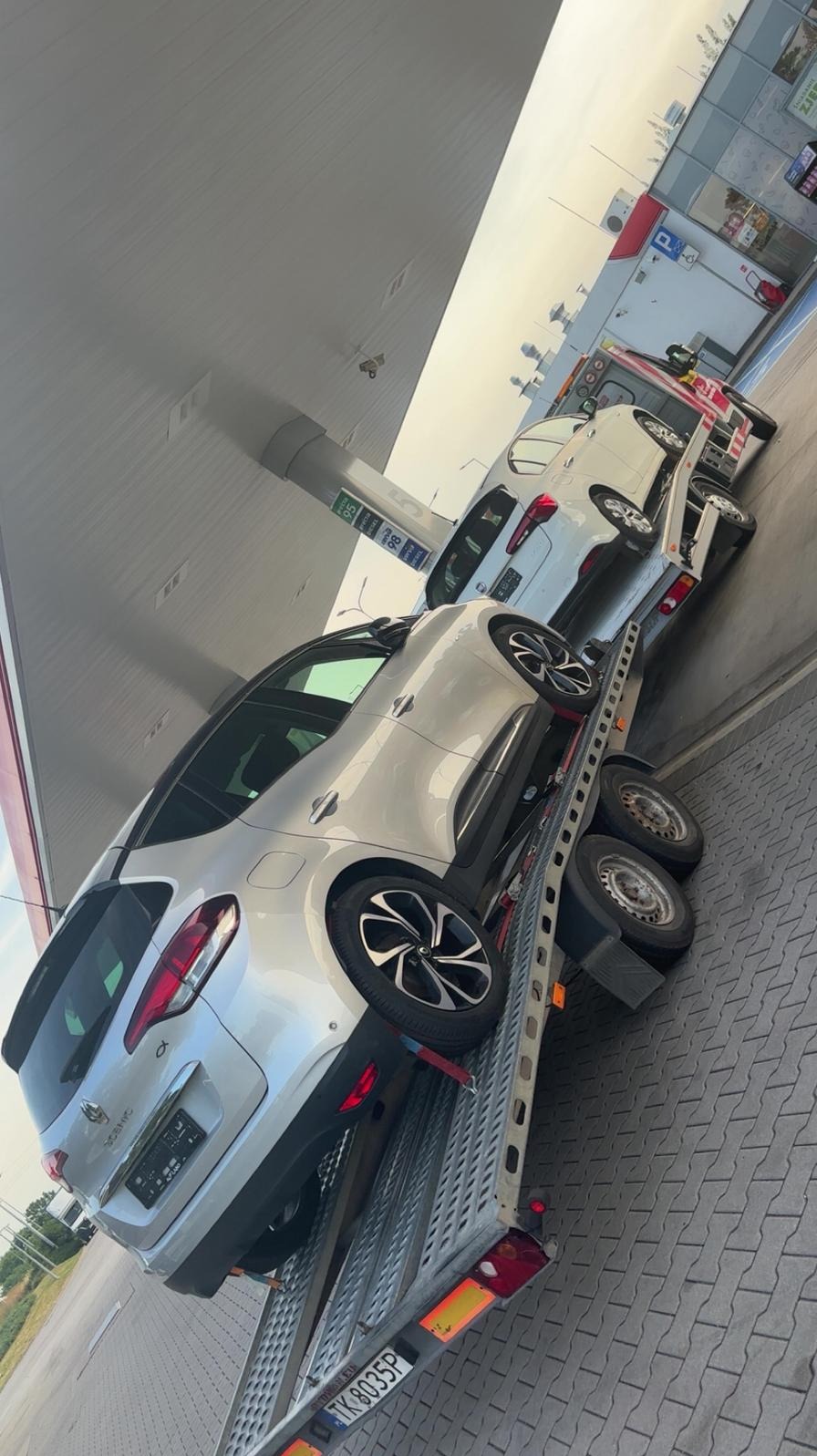 Dwa samochody osobowe, srebrny i biały, na lawecie pod zadaszeniem stacji benzynowej. Widoczny fragment tablicy rejestracyjnej lawety. Ujęcie z boku.