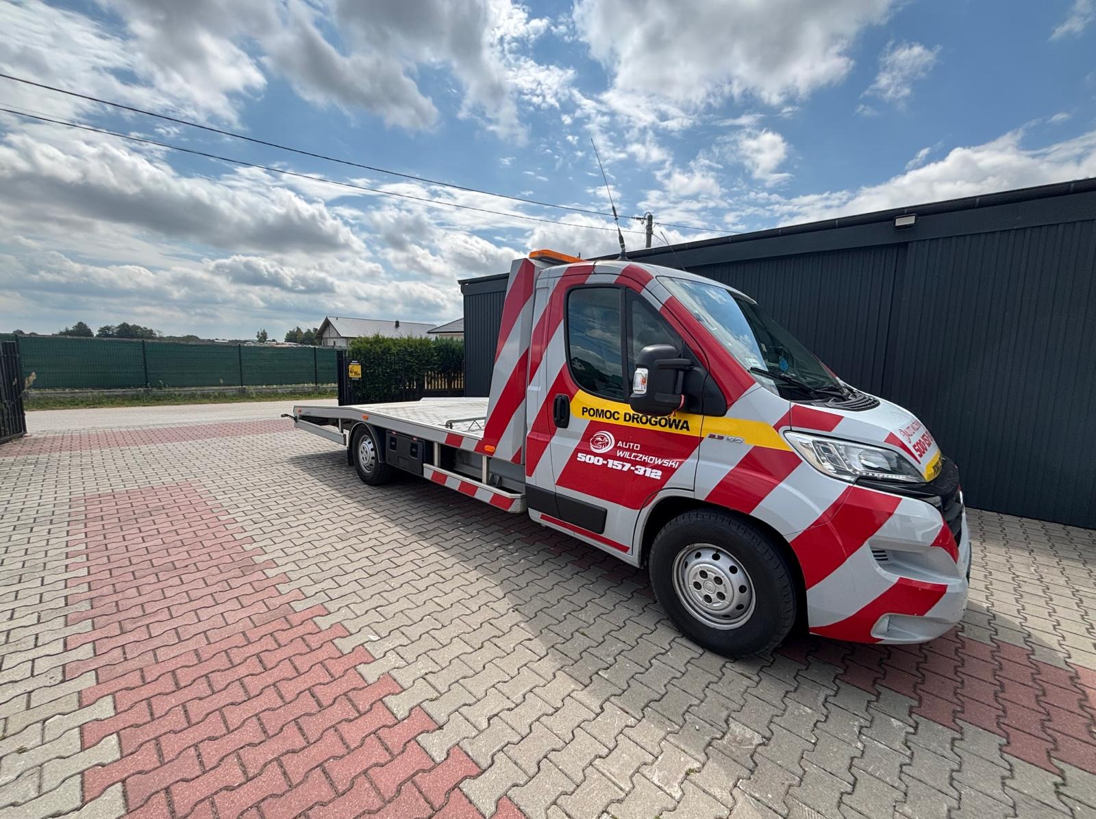 Czerwono-biała laweta z napisem 'Pomoc Drogowa' na tle kostki brukowej i pochmurnego nieba. Widoczny numer telefonu i logo firmy.