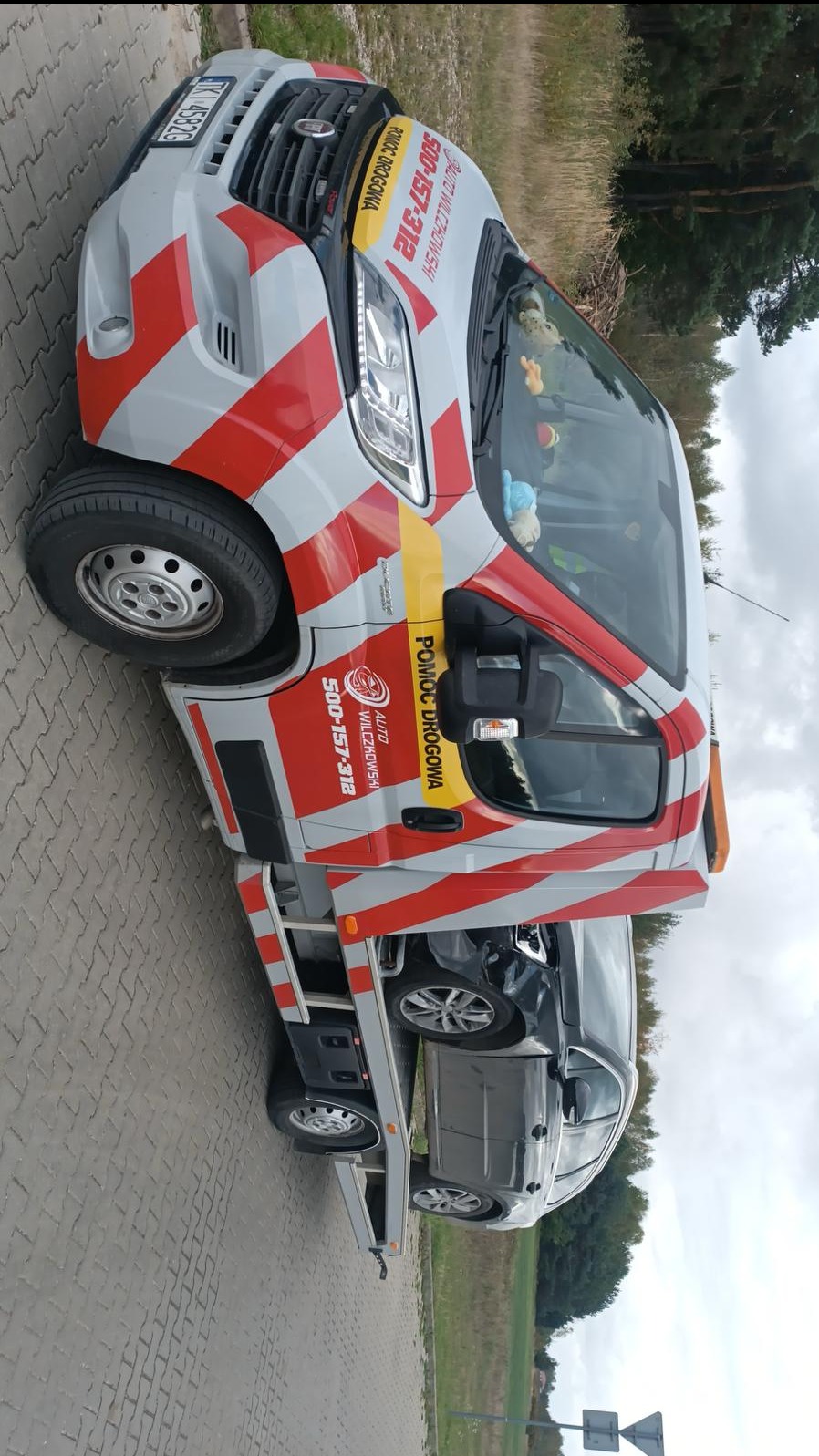 Laweta z uszkodzonym, szarym samochodem osobowym na platformie. Widoczny numer telefonu do pomocy drogowej i pluszowe zabawki w kabinie. Ujęcie z boku, na zewnątrz.
