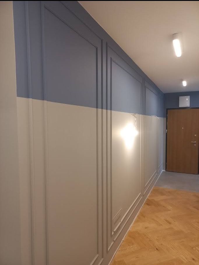 Korytarz po remoncie: ściany z dekoracyjnymi panelami w kolorze szaro-białym, drewniana podłoga w jodełkę, oświetlenie sufitowe i drzwi wejściowe w tle.