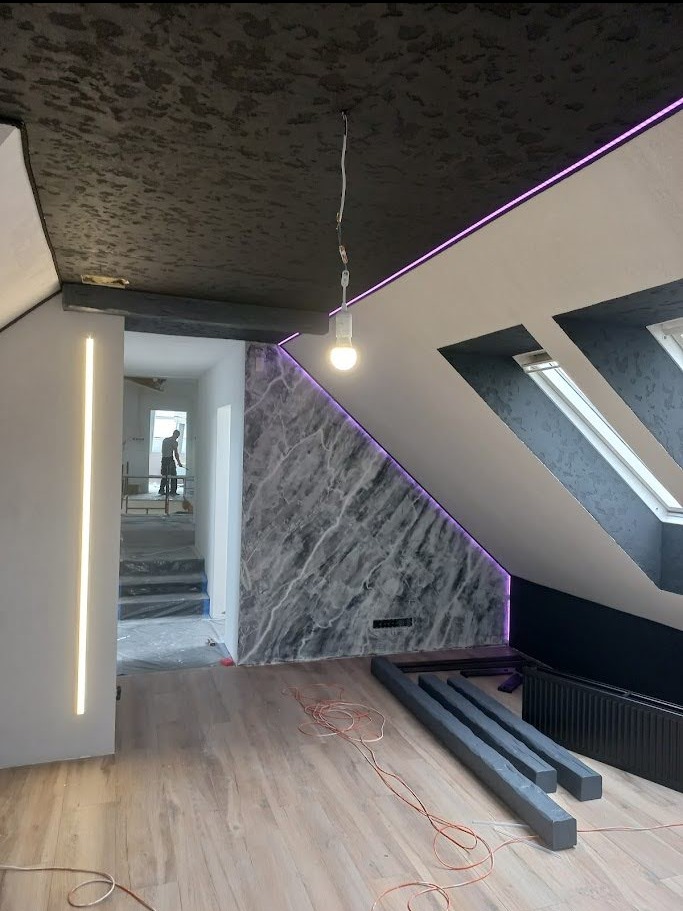 Wykończone poddasze ze ścianą imitującą marmur, oświetleniem LED i ciemnym, teksturowanym sufitem. W tle widoczny pracownik na schodach.