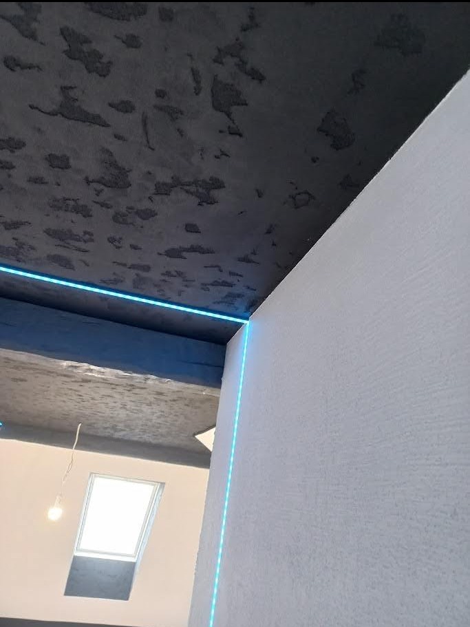 Szara, teksturowana powierzchnia sufitu z subtelnym oświetleniem LED, kontrastująca z gładką, białą ścianą. Widoczny fragment okna dachowego i wisząca żarówka.