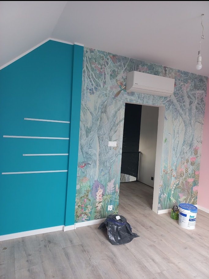 Pokój po malowaniu z turkusową ścianą i tapetą w leśny motyw. Widoczny klimatyzator, podłoga z paneli i akcesoria malarskie.