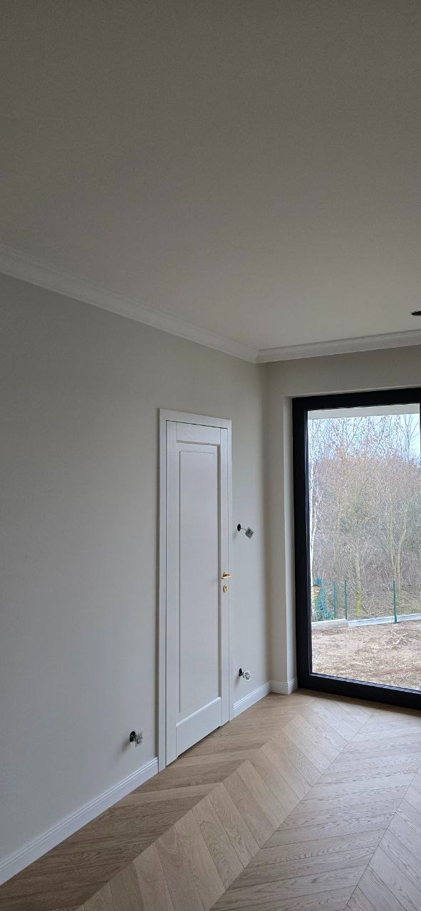 Jasne wnętrze z parkietem w jodełkę, białe ściany z listwami, drzwi i duże okno z widokiem na zewnątrz. Widoczne niedokończone gniazdka elektryczne.