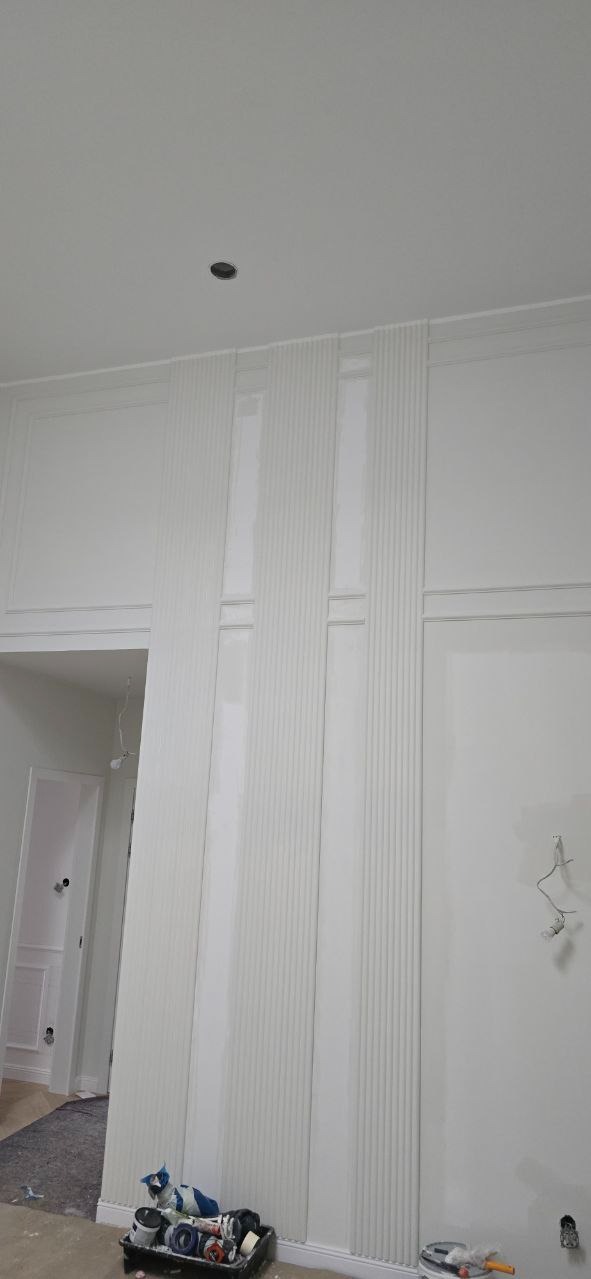 Białe panele dekoracyjne z pionowymi żłobieniami na ścianie, widoczne narzędzia malarskie, sufit z oświetleniem punktowym, wnętrze w trakcie wykańczania.