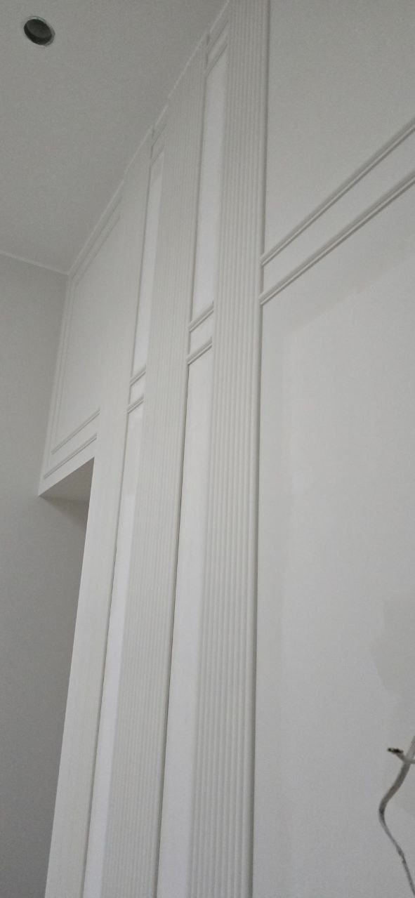 Białe, dekoracyjne panele ścienne z subtelnymi żłobieniami i listwami, tworzące elegancki wzór na ścianie. Minimalistyczny design, jasne oświetlenie.