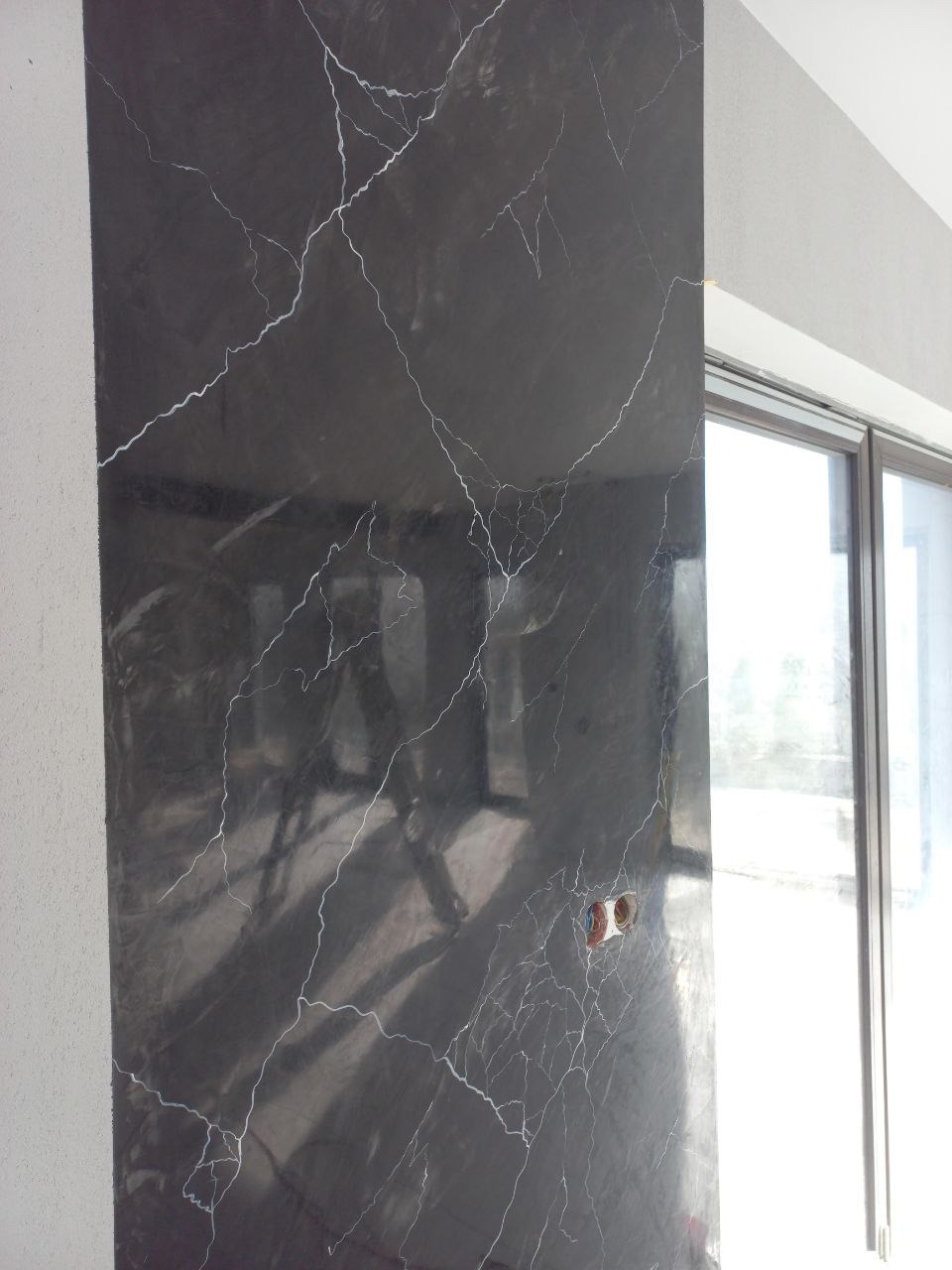 Ściana z efektem marmuru w odcieniach szarości, z widocznym gniazdkiem elektrycznym i fragmentem okna. Dekoracyjne wykończenie wnętrza.