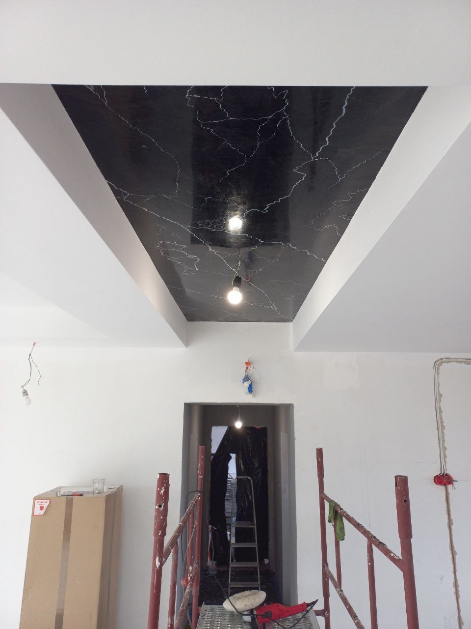 Czarny sufit z efektem marmuru i białymi obramowaniami w trakcie prac wykończeniowych. Widoczne rusztowania i narzędzia, sufit z oświetleniem punktowym.