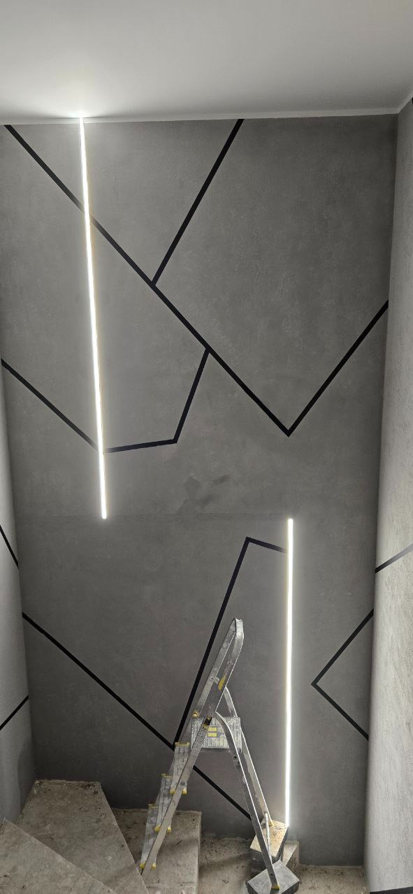 Szara ściana z geometrycznym wzorem z czarnych linii i pionowymi lampami LED. Na schodach stoi drabina.