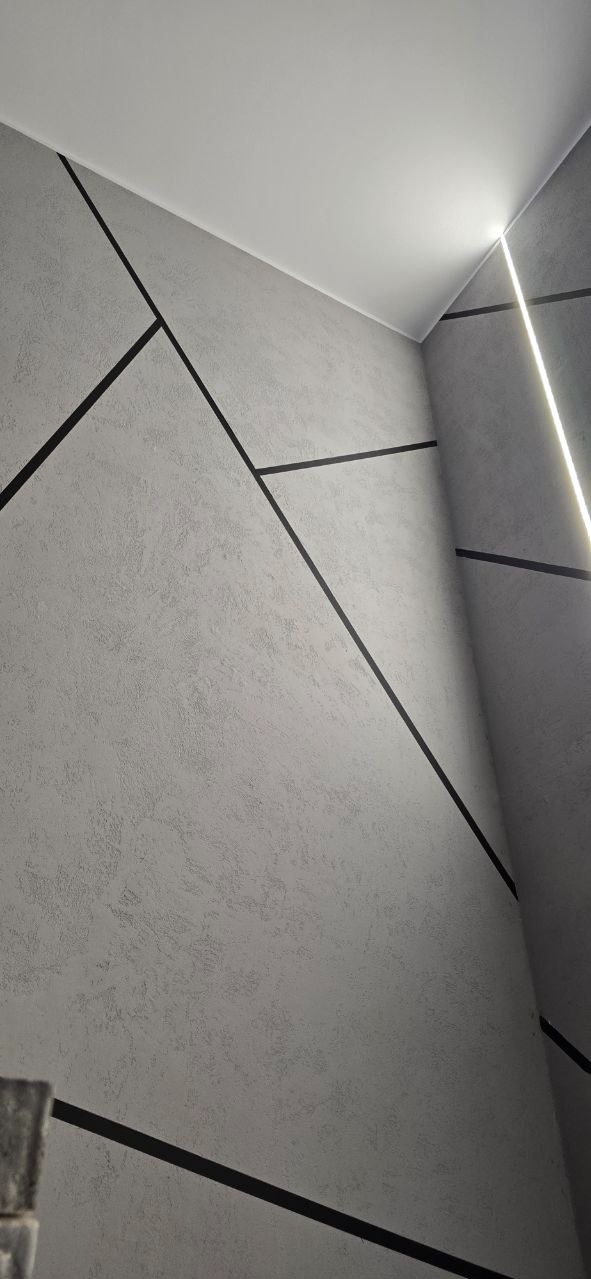 Szara ściana z dekoracyjnymi, czarnymi liniami tworzącymi geometryczny wzór. Oświetlenie LED w rogu sufitu. Teksturowana powierzchnia ściany.