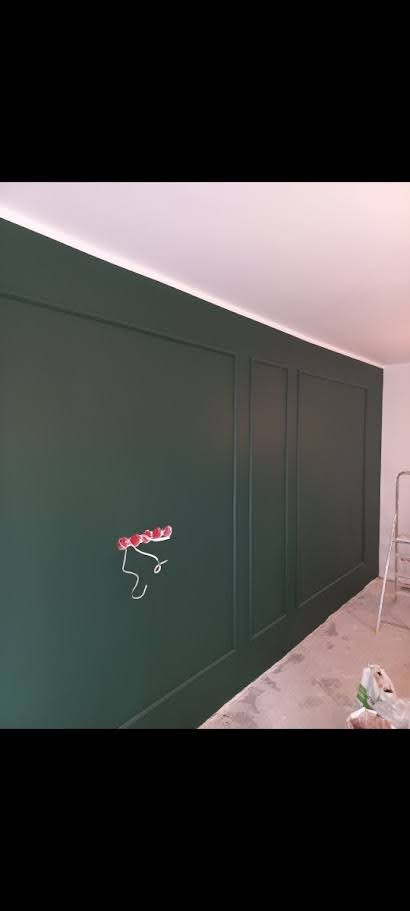 Elegancka, ciemnozielona ściana z dekoracyjnymi panelami i wystającą puszką elektryczną. Widoczna drabina i resztki po remoncie. Nowoczesne wykończenie wnętrza.