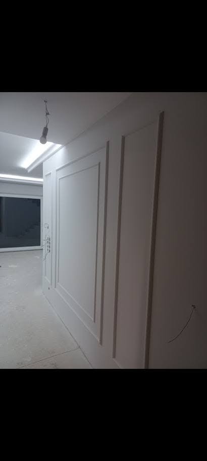 Biała ściana z dekoracyjnymi panelami ściennymi w stylu klasycznym. Widoczne oświetlenie sufitowe i fragment okna. Minimalistyczne wykończenie wnętrza.