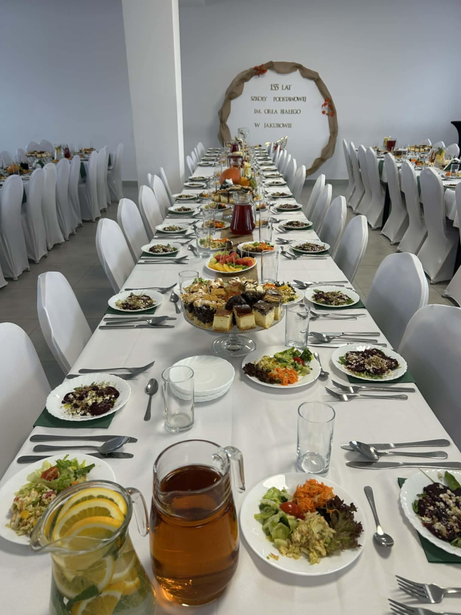 Uroczysty stół cateringowy z białym obrusem, zastawą i dekoracjami, przygotowany na 155-lecie szkoły podstawowej w Jakubowie. Widoczne sałatki, ciasta i napoje.