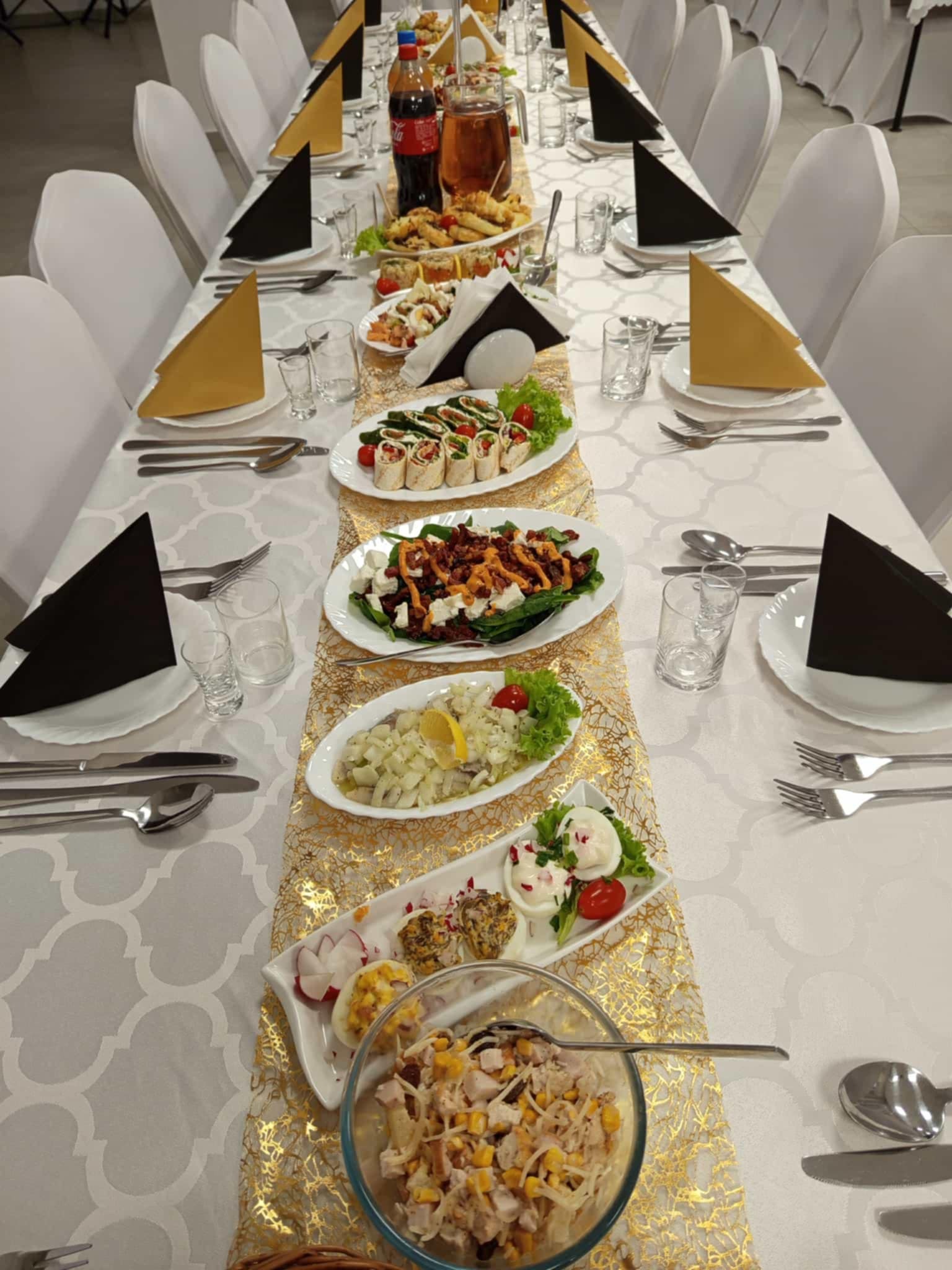 Uroczysty stół z białym obrusem, złotym bieżnikiem i elegancko ułożonymi potrawami, nakryciami i dekoracjami w odcieniach czerni i złota.