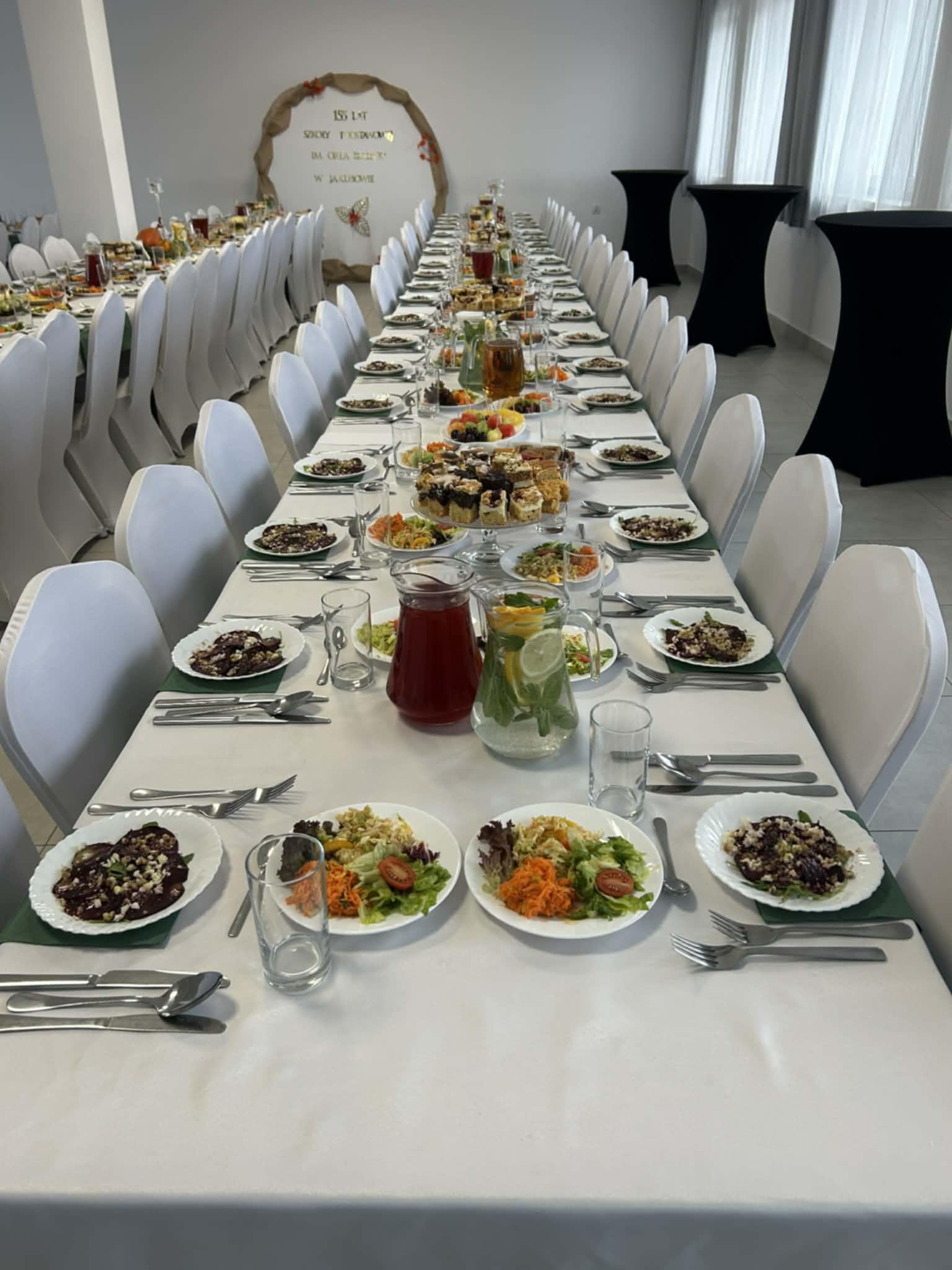 Długi stół z białym obrusem, nakryty dla gości z różnorodnymi daniami, napojami i sztućcami. Białe krzesła i dekoracja w tle sugerują uroczystość.