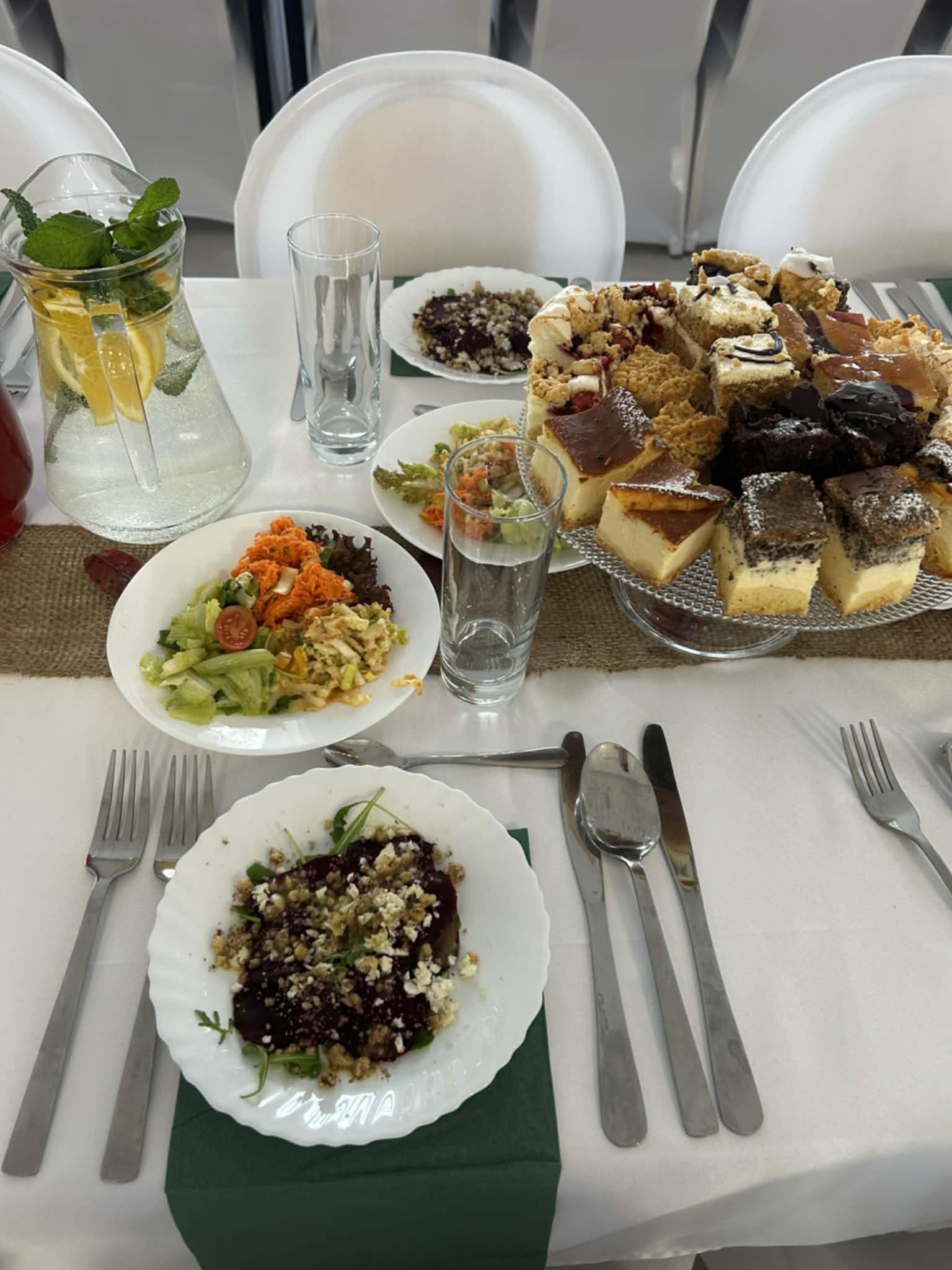 Stół z zastawą, sałatkami i paterą ciast. Dzbanek z wodą i cytryną. Nakrycie stołu w neutralnych barwach, z zielonym akcentem. Różnorodność dań na przyjęciu.