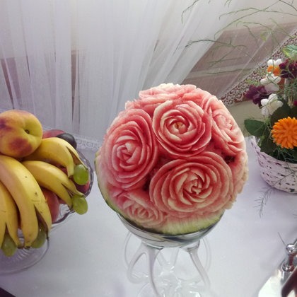 Arbuz rzeźbiony w róże na szklanej podstawie, obok owoce w paterze i kompozycja kwiatowa. Dekoracja stołu na przyjęciu.