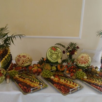 Dekoracyjny stół z owocami: rzeźbione arbuzy (róża, serce z gołębiami), ananas z owocami, tace z ułożonymi kolorystycznie owocami. Elegancka kompozycja na przyjęcie.