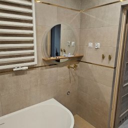 Usługi remontowo budowlane Przemysław Kozakiewicz - Nowoczesna łazienka w Wielgiem z wanną wolnostojącą, okrągłym lustrem z półką i grzejnikiem drabinkowym. Beżowe płytki z złotymi akcentami.