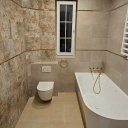 Usługi remontowo budowlane Przemysław Kozakiewicz - Elegancka łazienka z beżowymi płytkami, złotymi akcentami i nowoczesną armaturą. Uwagę przykuwa asymetryczna wanna i podświetlany sufit.