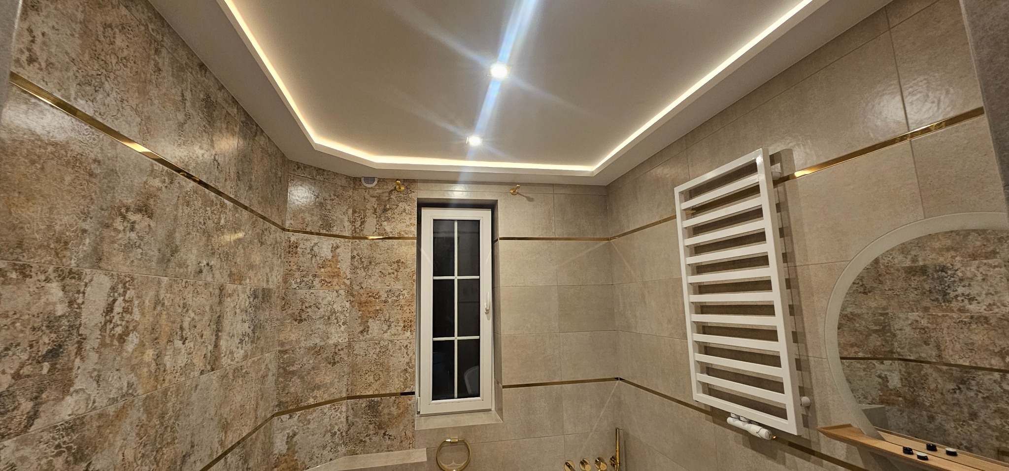 Elegancka łazienka z beżowymi płytkami, złotymi akcentami i oświetleniem LED. Widoczne okno, grzejnik drabinkowy i okrągłe lustro. Całość tworzy spójny, luksusowy wygląd.