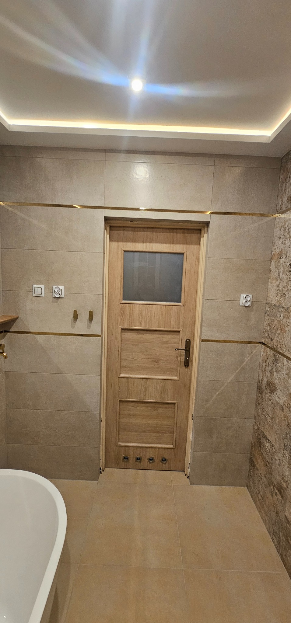 Jasna łazienka z beżowymi płytkami, drewnianymi drzwiami z szybą i złotymi akcentami. Widoczna część wanny i oświetlenie sufitowe LED.