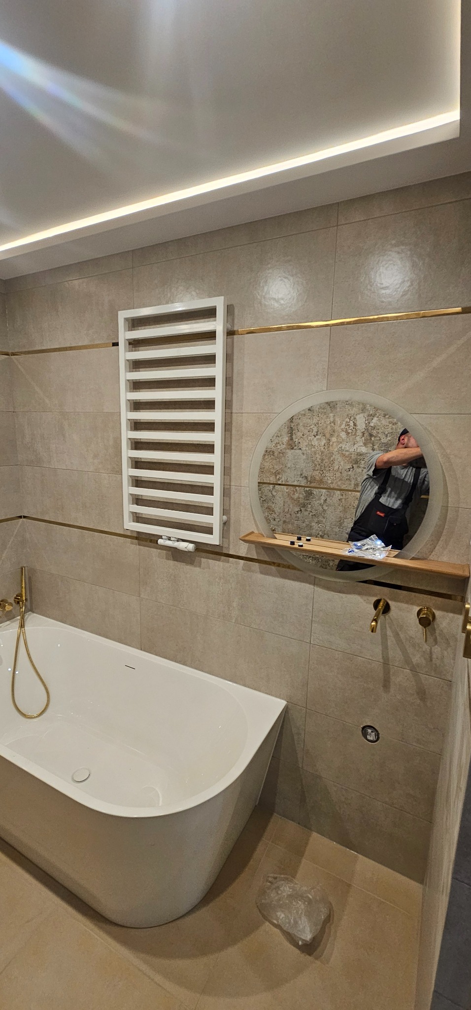 Nowoczesna łazienka z wanną, grzejnikiem drabinkowym i okrągłym lustrem, w którym odbija się instalator podczas montażu armatury. Beżowe płytki z złotymi akcentami.