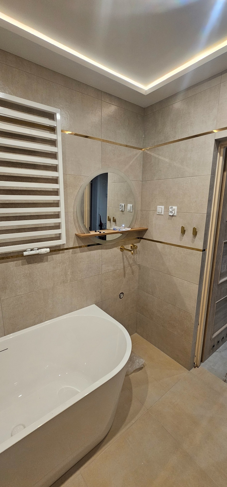 Nowoczesna łazienka w Wielgiem z wanną wolnostojącą, okrągłym lustrem z półką i grzejnikiem drabinkowym. Beżowe płytki z złotymi akcentami.