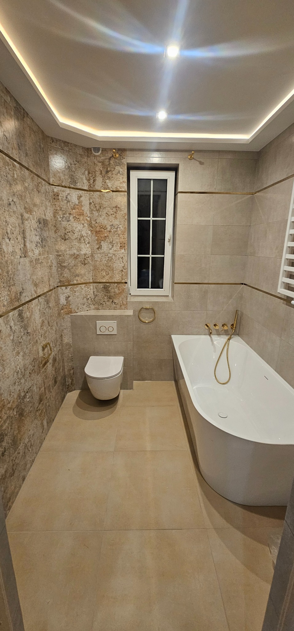 Elegancka łazienka z beżowymi płytkami, złotymi akcentami i nowoczesną armaturą. Uwagę przykuwa asymetryczna wanna i podświetlany sufit.