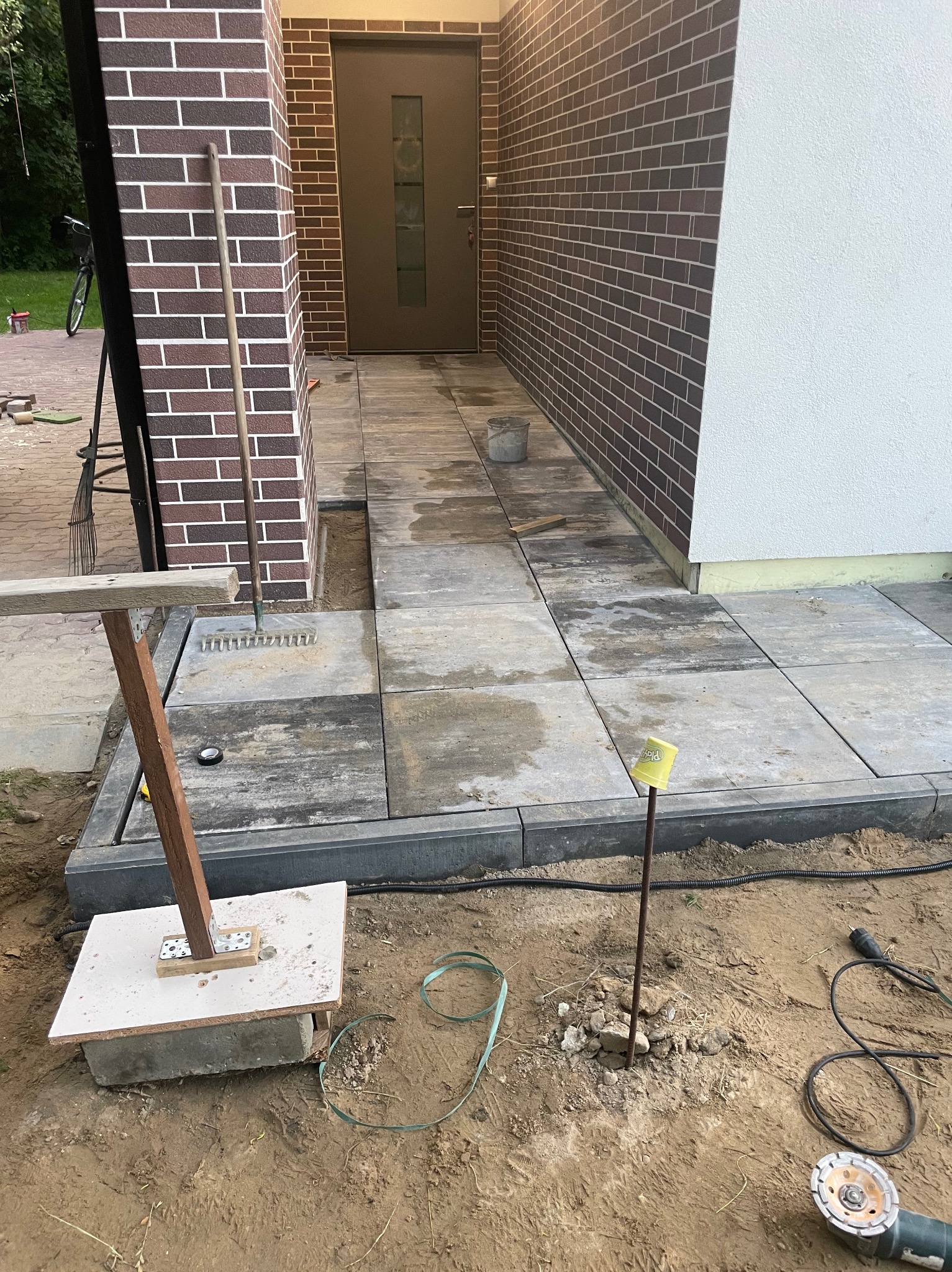 Układanie kostki brukowej na posesji. Widoczne narzędzia, fragment elewacji budynku z cegły i biały tynk. Prace w toku, teren przygotowany pod dalsze prace.