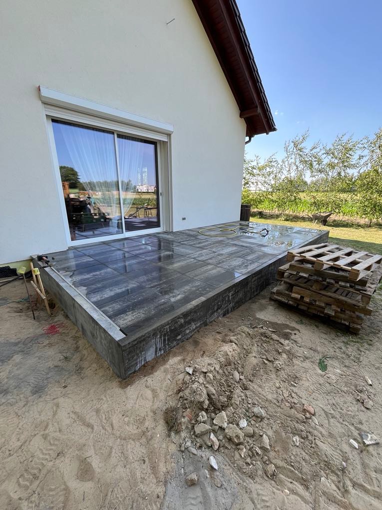 Nowy taras z ciemnych płyt przy domu z przesuwnymi drzwiami. Widoczne palety i piasek. Świeżo ułożona nawierzchnia, jeszcze mokra. Prace wykończeniowe w toku.
