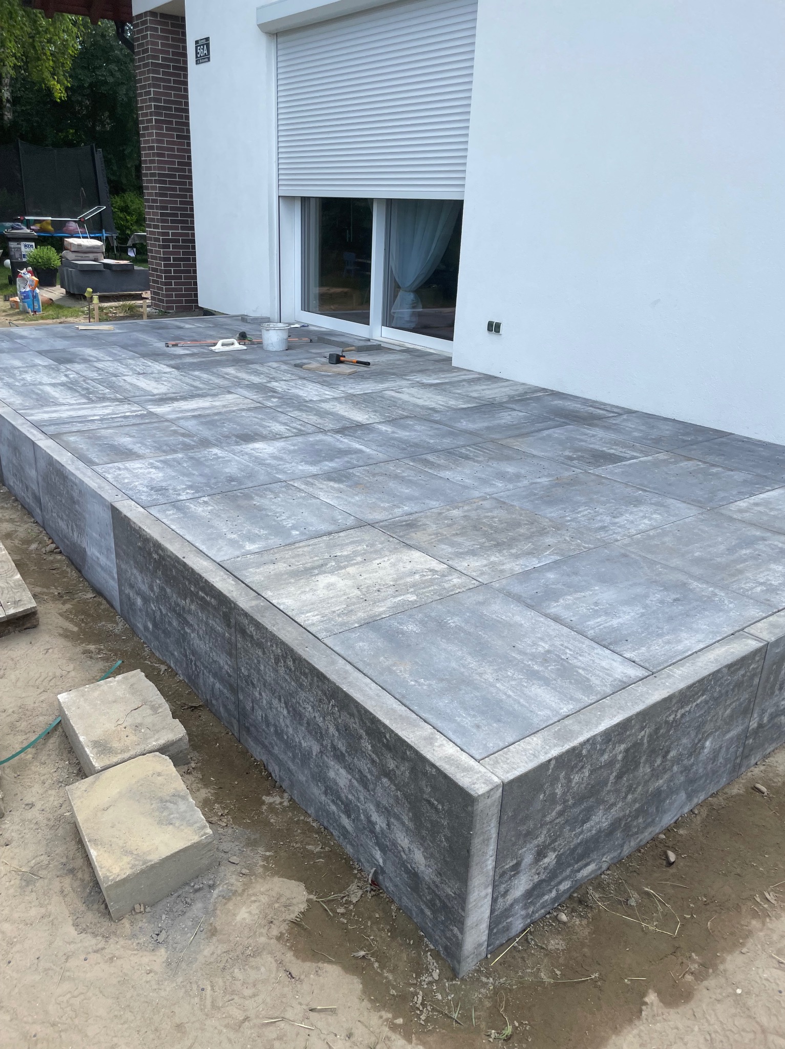 Nowoczesny taras z szarych płyt betonowych, częściowo ukończony, z widocznymi narzędziami i elementami wykończeniowymi. Minimalistyczny design, jasne tło budynku.