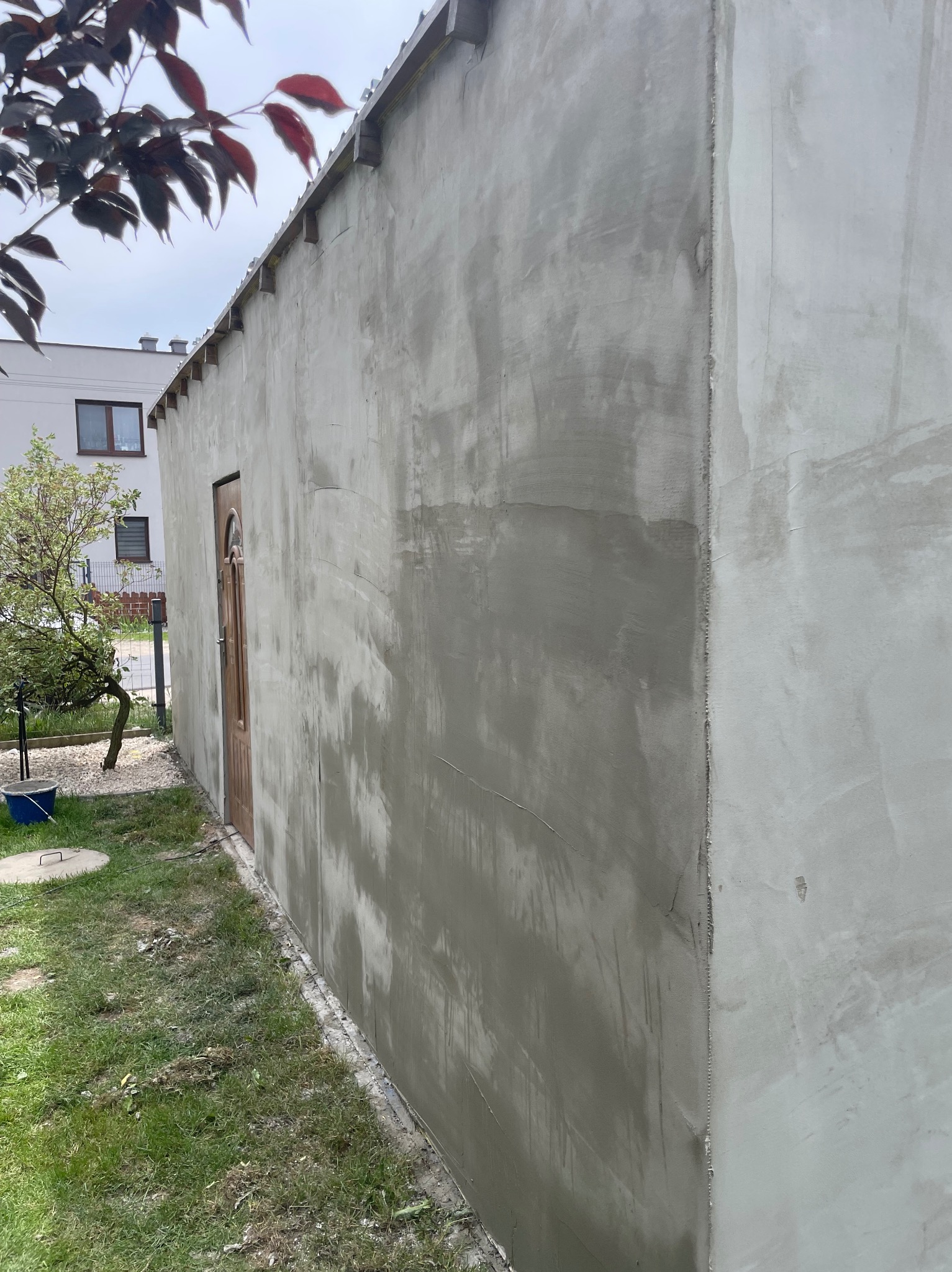Ściana budynku po nałożeniu szarej, surowej gładzi, widoczne łączenia płyt. Na ścianie drewniane elementy konstrukcyjne. Obok drzwi wejściowe i trawnik.