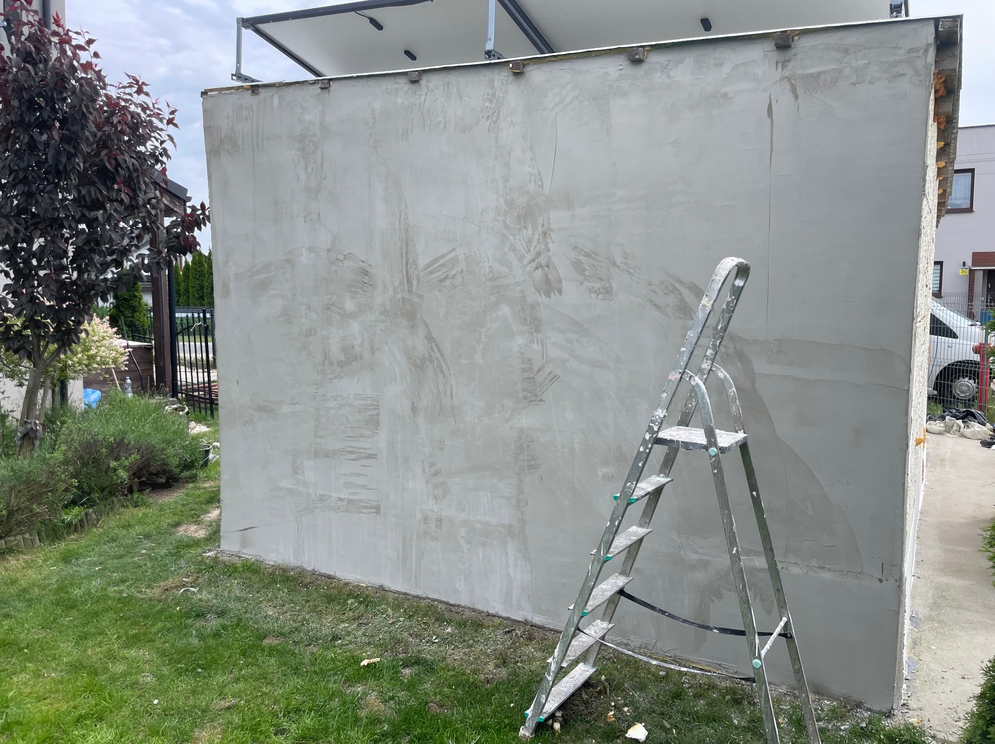 Ściana budynku w trakcie gładzenia, widoczne ślady po szpachli. Aluminiowa drabina oparta o ścianę. Trawa i roślinność w tle. Etap prac wykończeniowych na zewnątrz.