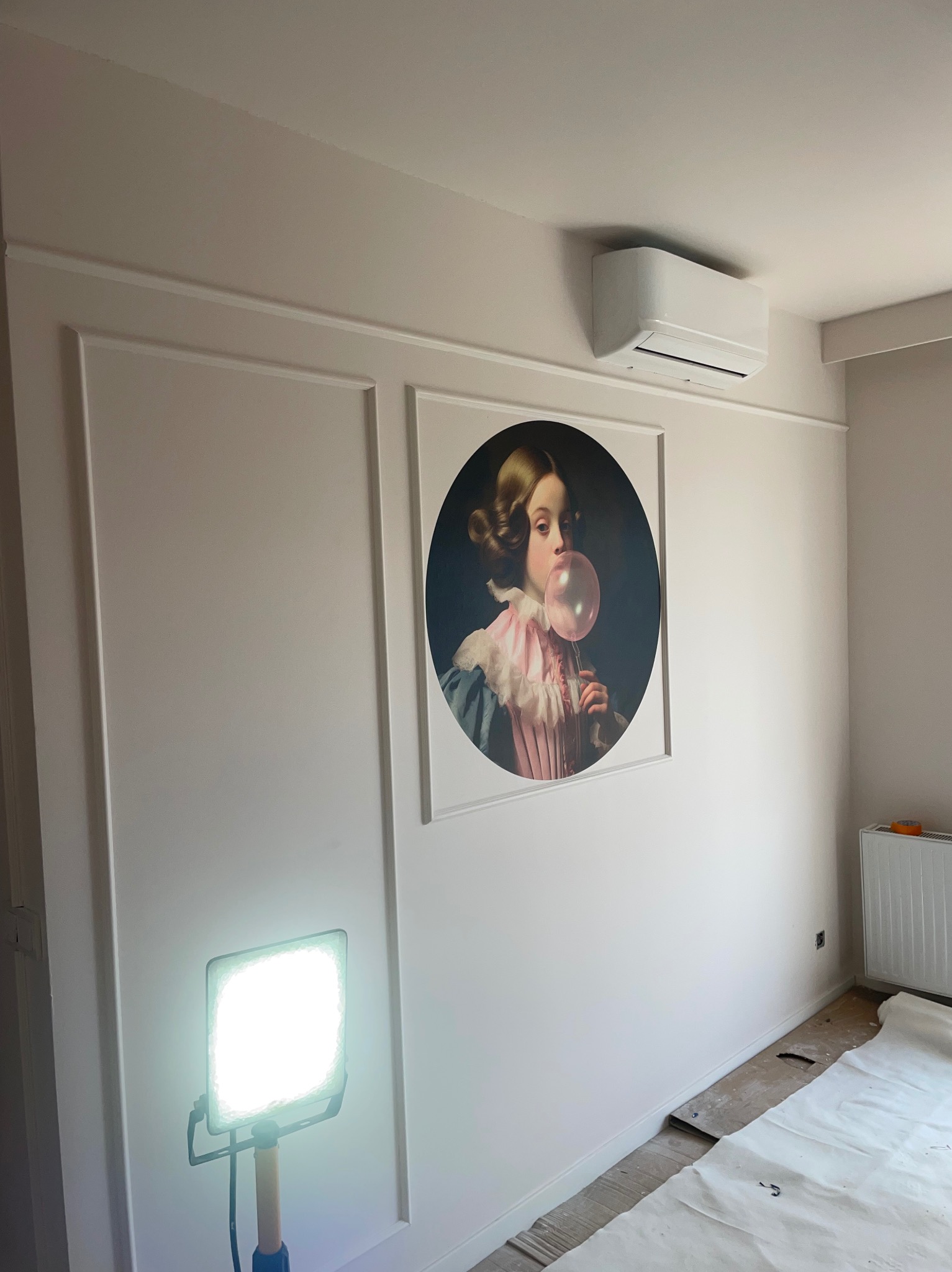 Wykończone wnętrze pokoju z dekoracyjnymi panelami ściennymi, obrazem w ramie i klimatyzatorem. Podłoga zabezpieczona folią malarską, widoczna lampa robocza.