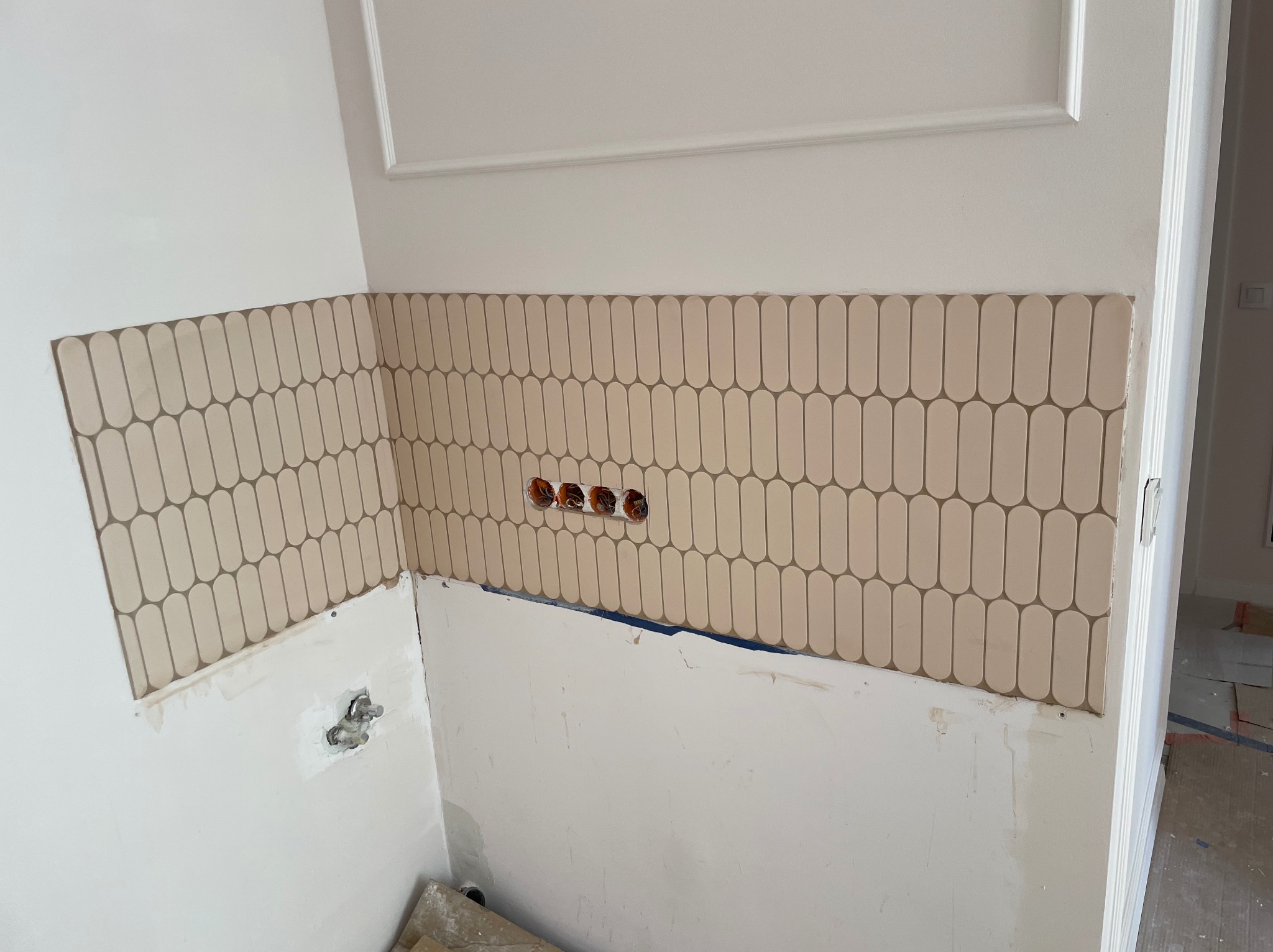 Fragment ściany z beżowymi, podłużnymi płytkami ułożonymi w pionie, nad niezakończonym fragmentem ściany. Widoczne gniazdka elektryczne i listwa dekoracyjna.