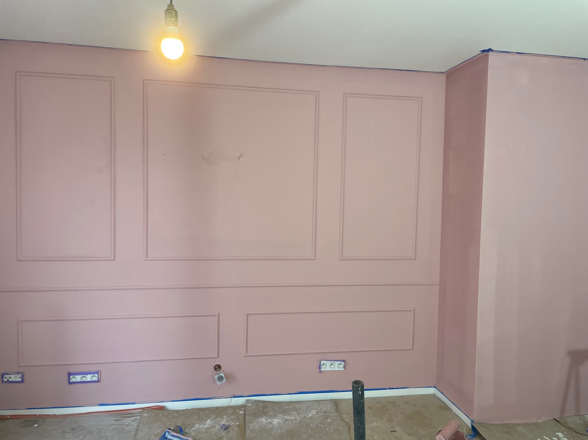Różowa ściana z dekoracyjnymi listwami i gniazdkami, zabezpieczona taśmą malarską przy podłodze. Widoczna żarówka. Styl klasyczny, elegancki detal wykończenia wnętrza.